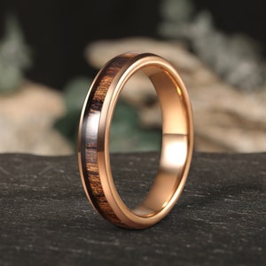 May include: A rose gold tungsten carbide ring with a brown wood inlay. The ring is polished and has a beveled edge.