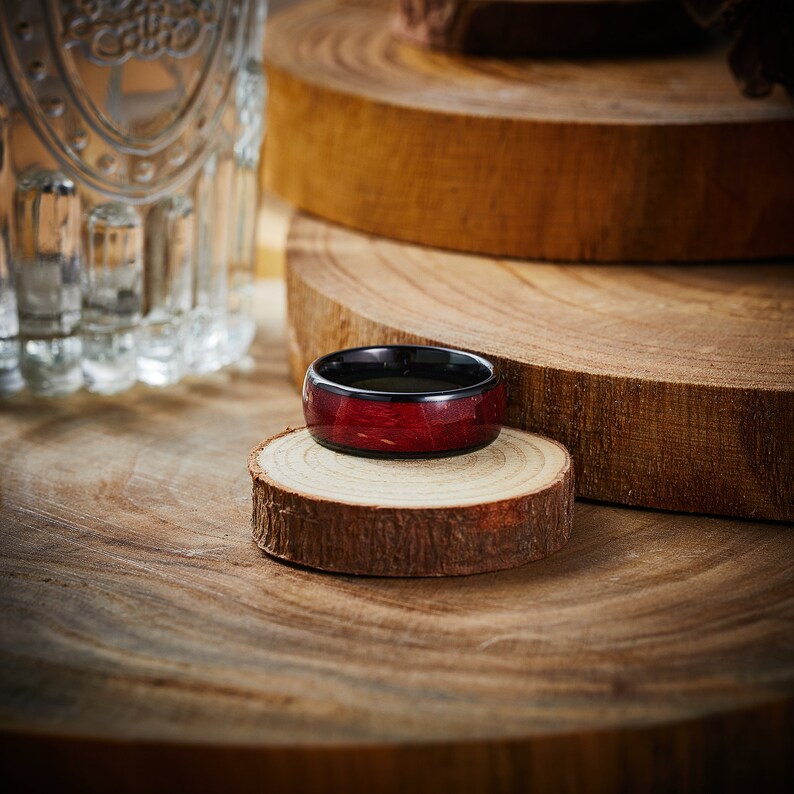 May include: A dark red and black wooden ring with a polished finish. The ring is sitting on a wooden surface.