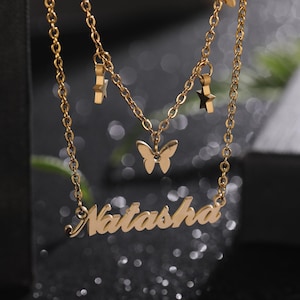 以下が含まれることがあります： 蝶のチャームと「Natasha」という名前が筆記体で書かれたゴールドトーンのネックレス。ネックレスには、ぶら下がった星のチャームが2つ付いています。