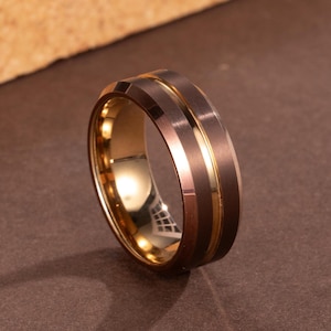 Puede incluir: Un anillo de dos tonos con un exterior marrón oscuro cepillado y un interior dorado. El anillo presenta una banda central dorada y un borde biselado. Es una joya.