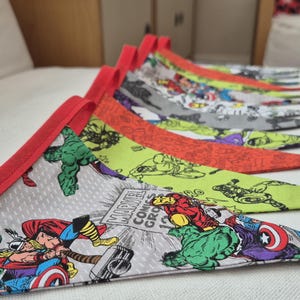 May include: A row of colorful triangular fabric banners with red trim. The banners feature comic book characters, including the Hulk, Thor, and Captain America. The fabric has a Marvel Comics Group logo.