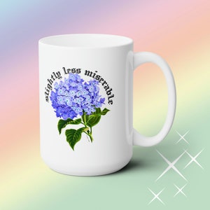 May include: White ceramic mug with a purple hydrangea flower graphic and the text "slightly less miserable" in black.