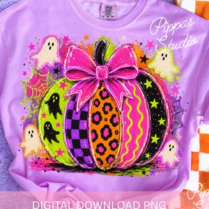 May include: A light purple t-shirt featuring a central pumpkin graphic with a large hot pink bow. The pumpkin has segments of checkerboard, leopard print, and wavy lines. Surrounding the pumpkin are small ghosts, stars, and spiderwebs.