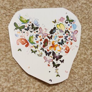 May include: A white heart-shaped sticker featuring a colourful design of butterflies. The butterflies are in various shades of blue, green, red, orange, yellow, and black. The butterflies are arranged in a heart shape.