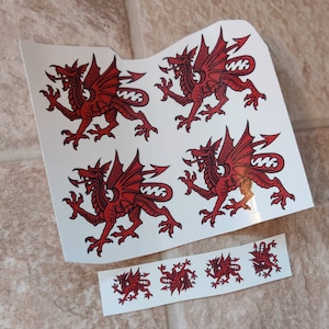 May include: Four red Welsh dragon decals on a white background. The dragons are facing different directions. There are five smaller versions of the dragon decals on a separate white sheet.