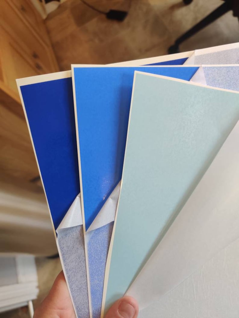 May include: Four sheets of adhesive paper in various shades of blue. The sheets are stacked on top of each other, with the top sheet being a light blue, the second sheet being a darker blue, and the bottom two sheets being a deep blue.