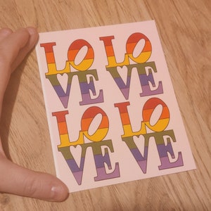 May include: A pink card with the word "LOVE" repeated four times in rainbow colours. Each letter is outlined in black and has a small heart in the centre.
