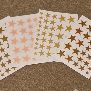 May include: Three sheets of white paper with gold star stickers. The stars are of different sizes and are arranged in a random pattern.