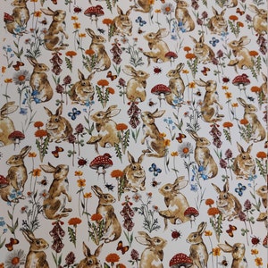 May include: A white fabric with a repeating pattern of brown rabbits, flowers, and mushrooms. The rabbits are in various poses, some sitting, some standing, and some hopping. The flowers are in shades of pink, purple, blue, and yellow. The mushrooms are red with white spots.