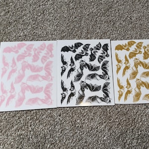 May include: Three sheets of paper with black and white bat illustrations. The bats are printed on a white background. The sheets are pink, black, and gold.
