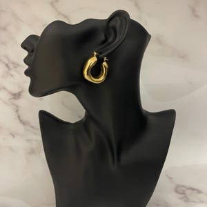 Op de afbeelding: Gouden creolen met een dik, afgerond ontwerp. De oorbellen worden getoond op een zwart mannequinhoofd.