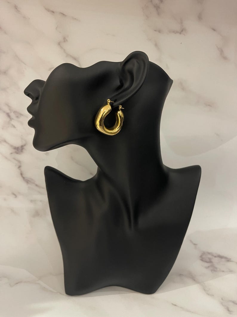 Op de afbeelding: Gouden creolen met een dik, afgerond ontwerp. De oorbellen worden getoond op een zwart mannequinhoofd.
