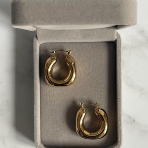 Op de afbeelding: Een paar gouden creolen in een grijze fluwelen doos. De oorbellen zijn dik en hebben een afgeronde, licht ovale vorm.