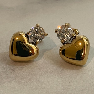 May include: A pair of gold-colored heart-shaped stud earrings. Each earring features a polished, rounded heart base with a sparkling, round, clear gemstone set on top. The earrings are displayed on a light-colored, marbled surface.