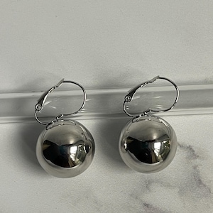 May include: A pair of shiny, silver ball earrings. Each earring features a large, spherical polished metal ball suspended from a lever-back clasp. The earrings reflect the surrounding light, creating a bright, metallic appearance. The jewelry is displayed against a white background.