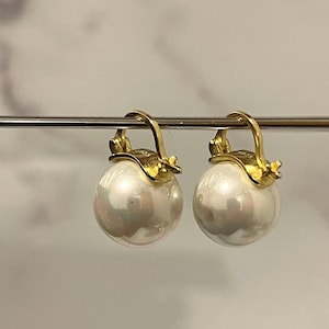 Puede incluir: Un par de pendientes de perlas con detalles dorados. Cada pendiente presenta una perla blanca redonda y grande suspendida de un cierre de palanca dorado. Los detalles dorados tienen un diseño floral.