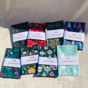 Op de afbeelding: Een verzameling gevouwen stoffen wraps, elk met een uniek patroon met een vakantiethema. Ontwerpen omvatten kerstbomen, ornamenten en bloemmotieven in groen, rood en blauw. Elke wrap is gelabeld met "Petit Furoshiki".