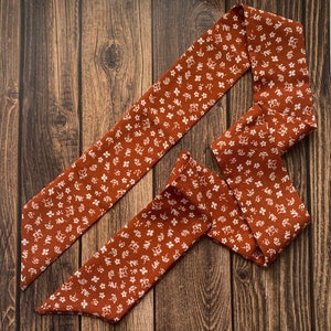 May include: A rust-colored fabric hair scarf with a white floral pattern. The scarf is long and narrow, designed to be tied around the head or neck. The floral design is composed of small, delicate white flowers.
