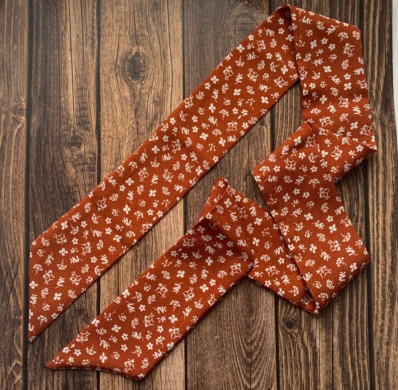 May include: A rust-colored fabric hair scarf with a white floral pattern. The scarf is long and narrow, designed to be tied around the head or neck. The floral design is composed of small, delicate white flowers.