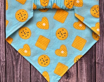Puede incluir: Bandana azul claro para mascotas con un patrón repetido de gofres y galletas amarillas. La bandana tiene una banda elástica. El diseño incluye formas de comida en forma de corazón, cuadradas y redondas.