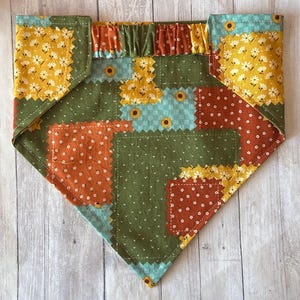 May include: A triangular pet bandana featuring a patchwork design of various floral and geometric patterns in shades of olive green, mustard yellow, burnt orange, and light blue. The top edge includes a gathered fabric channel for a collar.