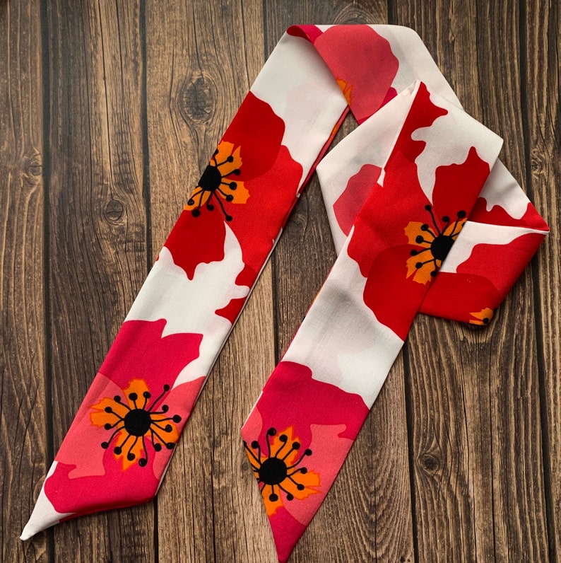 May include: A long, floral scarf featuring a vibrant red poppy design with black and orange centers against a white background. The scarf is made of a lightweight fabric and is displayed on a wooden surface.