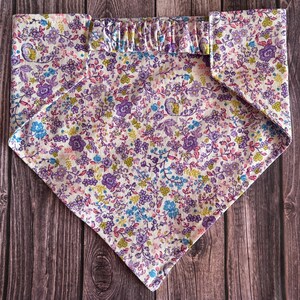 May include: A white bandana with a floral pattern in shades of purple, pink, blue, and green. The bandana has a ruffled edge.