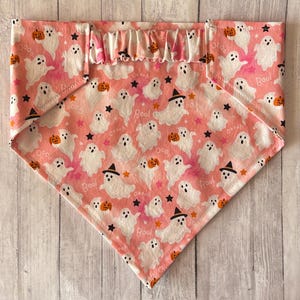 May include: A triangular pet bandana featuring a light pink background with a repeating pattern of white ghosts, orange pumpkins, black witch hats, and small black stars. The fabric includes the text Boo printed in a script font throughout.