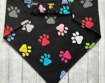 May include: Black bandana with a colorful paw print pattern. The bandana has a ruffled trim and is tied around the neck.