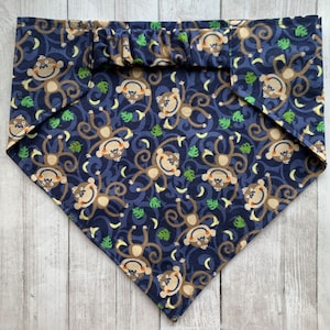 May include: A blue bandana with a pattern of brown monkeys and yellow bananas on a dark blue background. The bandana has a ruffled edge.
