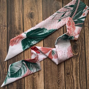May include: A white and pink floral patterned scarf with green leaves and accents of red and pink. The scarf is made of a soft material and is displayed on a wooden surface. The scarf is a fashion accessory.