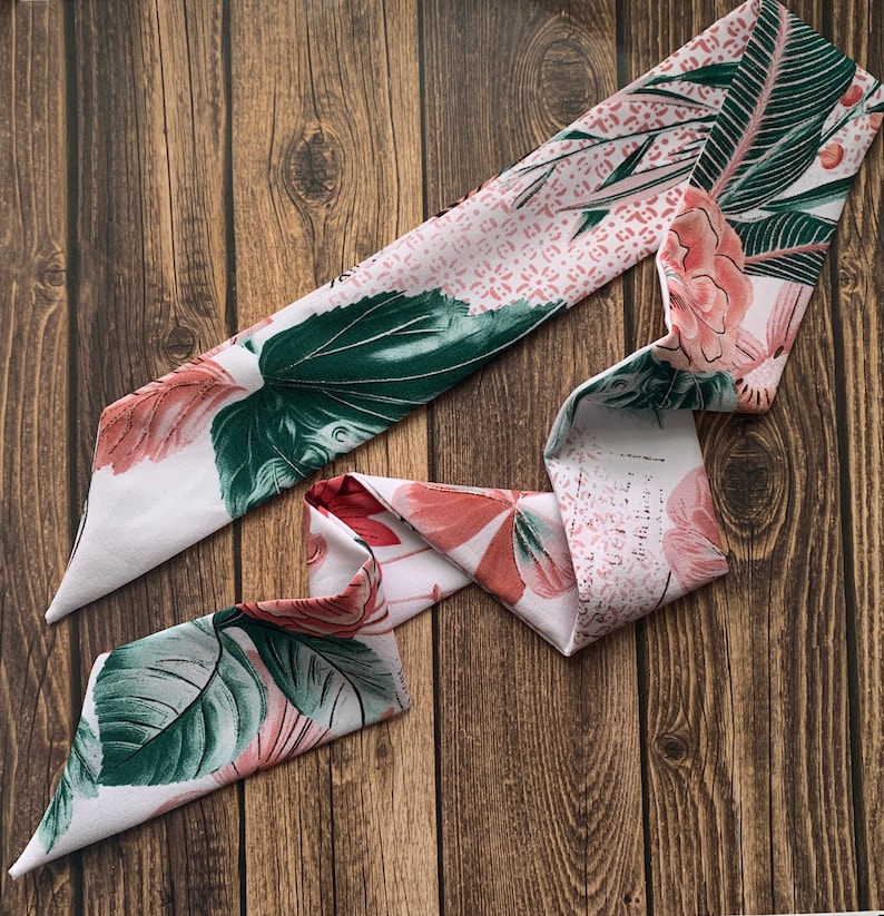 May include: A white and pink floral patterned scarf with green leaves and accents of red and pink. The scarf is made of a soft material and is displayed on a wooden surface. The scarf is a fashion accessory.