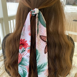 May include: A white hair scarf with a floral and butterfly design. The scarf features red flowers, green leaves, and a brown butterfly. The scarf is tied in a knot, with the ends hanging down.