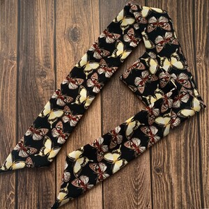 May include: A black hair scarf featuring a repeating pattern of butterflies in shades of white, yellow, and brown. The scarf is made of a soft, flowing fabric and is designed to be tied around the head or neck.