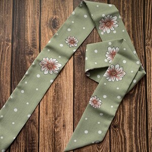 May include: A sage green scarf with a repeating pattern of white polka dots and large floral designs. The flowers have white petals and a brown center. The scarf is made of a soft fabric and is displayed on a wooden surface.
