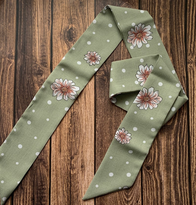 May include: A sage green scarf with a repeating pattern of white polka dots and large floral designs. The flowers have white petals and a brown center. The scarf is made of a soft fabric and is displayed on a wooden surface.