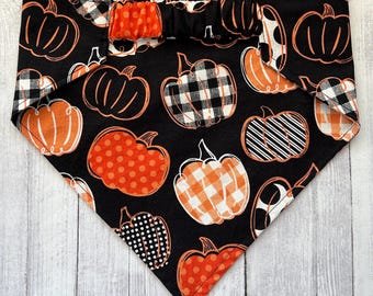 Peut inclure: Bandana noire pour chien avec un motif répété de citrouilles orange de différents motifs, dont des pois, des rayures et des carreaux. Le bandana a un nœud en haut et est posé sur un fond en bois blanc.