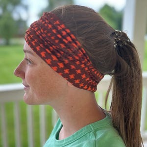 May include: A wide, stretchy fabric headband featuring a dark brown base color with a repeating, small-scale orange houndstooth pattern. The accessory is worn wrapped around the head, covering the hairline and ears with a gathered, textured appearance.