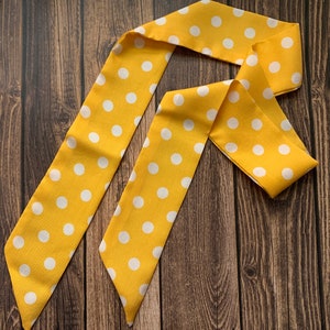 May include: A yellow polka dot scarf or hair accessory. The fabric is a bright yellow with evenly spaced white polka dots. The scarf is folded and arranged on a dark wood surface.
