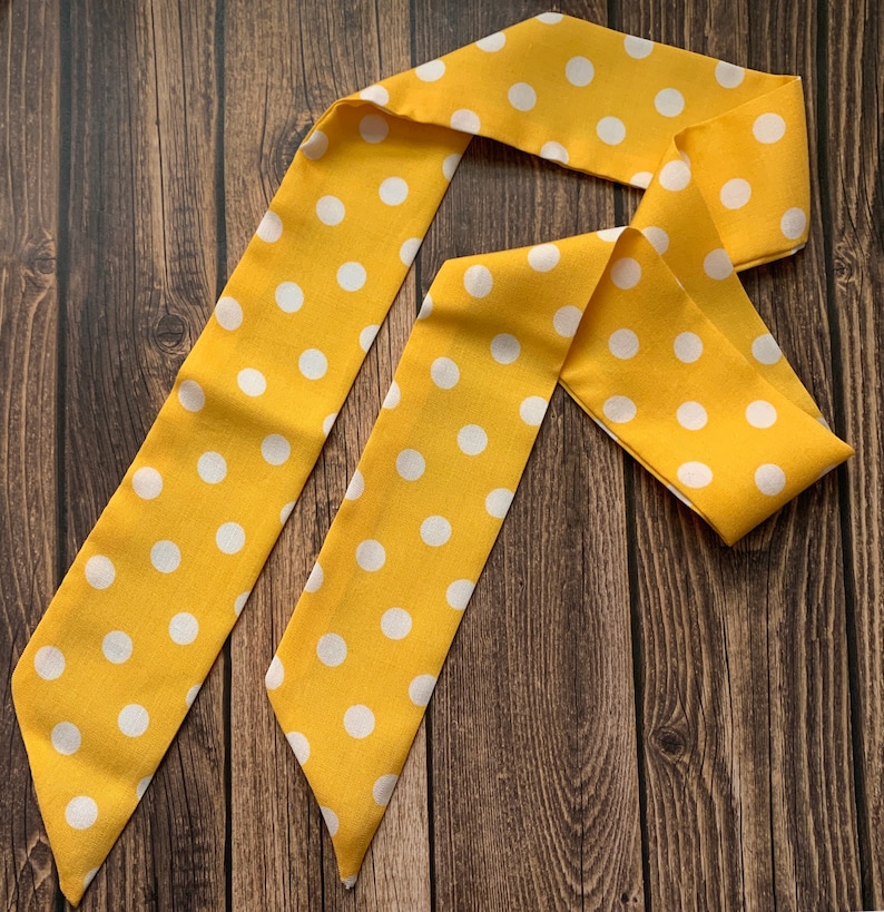 May include: A yellow polka dot scarf or hair accessory. The fabric is a bright yellow with evenly spaced white polka dots. The scarf is folded and arranged on a dark wood surface.