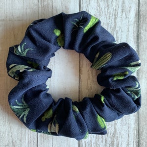 May include: A navy blue scrunchie with a cactus print. The scrunchie is made of a soft, stretchy fabric and is perfect for holding hair in place.