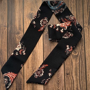 May include: A black scarf with a paisley pattern in shades of red, white, and blue. The scarf is made of a soft fabric and is folded in a decorative manner, showcasing the intricate floral design.