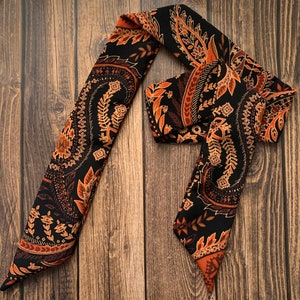 May include: A black and brown paisley patterned scarf. The scarf features a repeating design of floral and leaf motifs in shades of brown and orange against a black background. The scarf is made of a soft, flowing material.