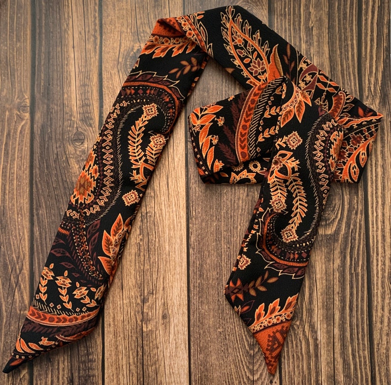 May include: A black and brown paisley patterned scarf. The scarf features a repeating design of floral and leaf motifs in shades of brown and orange against a black background. The scarf is made of a soft, flowing material.