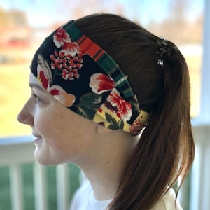 May include: A wide headband featuring a floral pattern in red, white, and yellow flowers on a black background. The headband also has a green and orange plaid design. The accessory is worn on the head.
