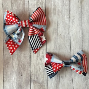 May include: Two patriotic fabric bows with red, white, and blue patterns. One bow features red stars on a white background, while the other has red and white stripes. The bows are on a light wood-grain surface.