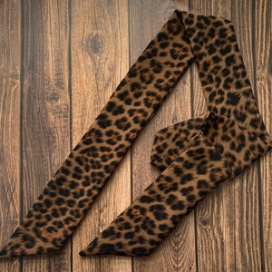 May include: A brown and black leopard print scarf is displayed on a wooden surface. The scarf is long and narrow, with a repeating pattern of dark brown spots on a lighter brown background. The scarf is folded and draped.