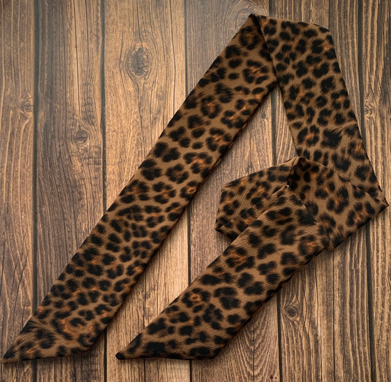May include: A brown and black leopard print scarf is displayed on a wooden surface. The scarf is long and narrow, with a repeating pattern of dark brown spots on a lighter brown background. The scarf is folded and draped.