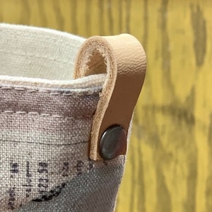 May include: A beige canvas tote bag with a brown leather handle. The bag has a printed design of abstract shapes in shades of blue, yellow, and brown. The text "BAL BARGAIN L", "ORTHWHILE", "FAMOUS QUALITY LINENS, BEDDING", "DRAPES AND MATERIALS", "TICALE SHEETS", and "CLOTHS" is printed on the bag.