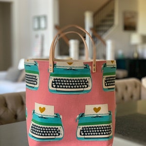 May include: A pink canvas tote bag with a typewriter design. The typewriter is blue, green, and white with a gold heart on the paper. The bag has brown leather handles.